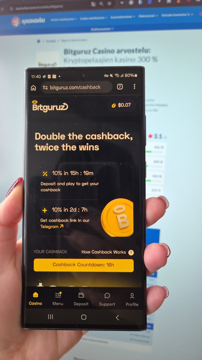 Käsi pitelee puhelinta, jonka näytöllä on Bitguruzin cashback-tarjous, ja taustalla näkyy tietokoneella kasinoarvostelu.