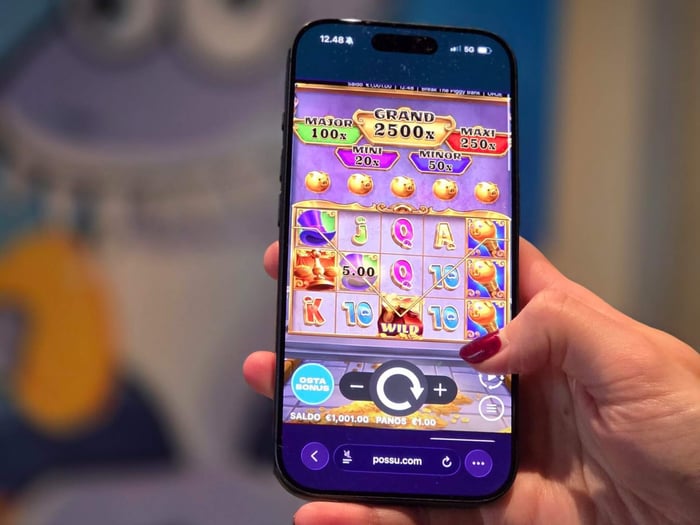 Käsi pitelee älypuhelinta, jonka näytöllä pyörii Possu Casinon värikäs kolikkopeli mobiiliversiossa, näyttäen voittokertoimet ja pelisaldon näytön alareunassa.