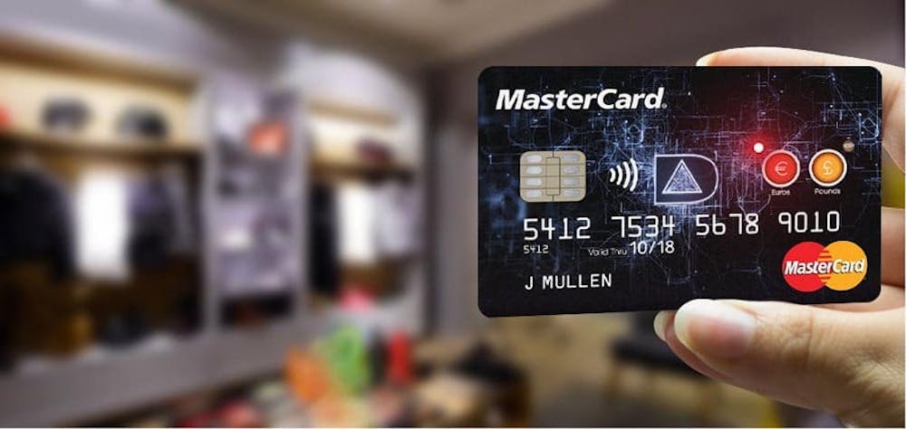 Käsi ja Mastercard-kotti etualalla, sumea tausta. Mastercard-kortissa on omaperäinen musta tausta, kortin logo, siru, numero ja kortinhaltijan nimi.